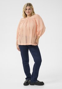 Peach-colored blouse with eyelet detailing, puffed sleeves, and a relaxed fit, paired with dark blue jeans and black loafers.