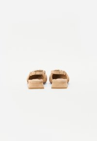 Sandales glissantes en suède beige avec un talon bloc bas, présentant une quincaillerie décorative sur le dessus et une surface lisse et texturée.