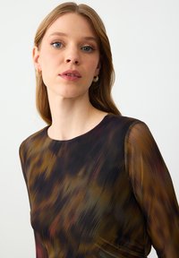 Jonge vrouw met lichtbruin haar, draagt gouden creolen en een donker, multikleurig patroon doorzichtig langmouwtop tegen een effen achtergrond.