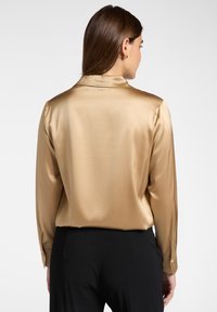 Frau in einer goldenen satinlangen Bluse mit langen Ärmeln und schwarzen Hosen, von hinten vor einem schlichten weißen Hintergrund gezeigt.