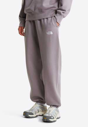 Pantalones deportivos grises con cintura elástica y bolsillos laterales, con un logo blanco en la parte superior del muslo izquierdo. Combinados con zapatillas grises y beige.