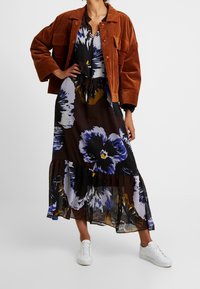 Brun kordjacka över en blommig, veckad maxikjol med blå, gula och vita nyanser; tillsammans med vita sneakers.