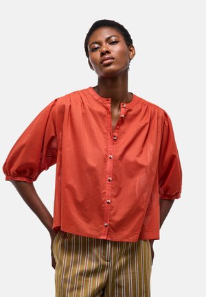 Femme aux cheveux courts portant un chemisier orange ample boutonné avec des manches bouffantes et un pantalon rayé marron, debout les mains sur les hanches.