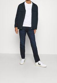 Mörkblå zip-up jacka med två framfickor över en vit t-shirt. Bärs med mörka jeans och vita sneakers med svarta detaljer.