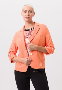 Koraalblazer met een getailleerd ontwerp, twee knopen en omgeslagen mouwen. G gedragen over een gestreept shirt en zwarte aansluitende broek met tekstdetails.