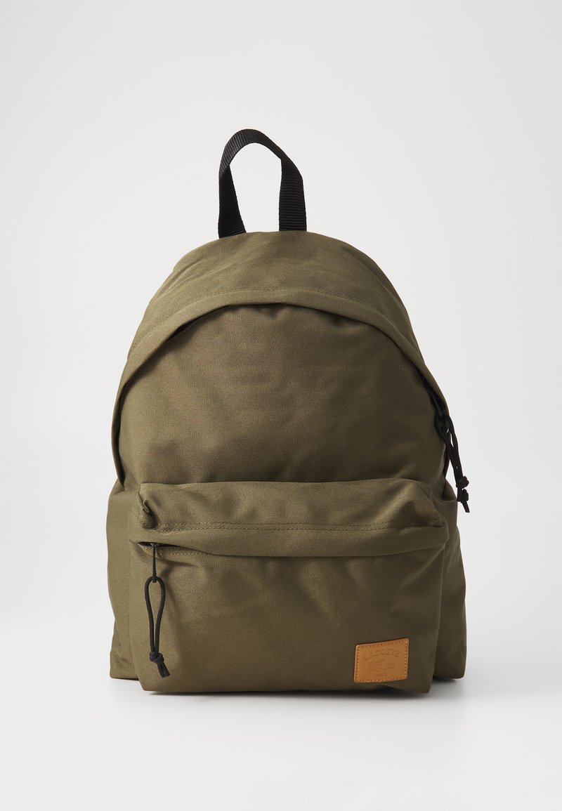 Mochila de tecido verde-oliva com forma arredondada, com um bolso frontal com fecho e um emblema de pele texturizada. Apresenta alças e detalhes em nylon preto.