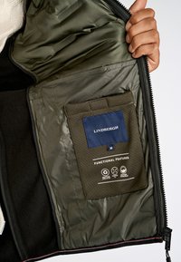 Svart jackas inredning med grön foder, en blå etikett med "LINDBERGH" och fickdetaljer som visar funktioner.