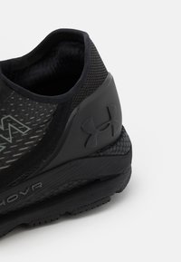 Zapatilla deportiva negra con parte superior de malla texturizada, cuello acolchado y suela de goma. Presenta un prominente logo de Under Armour en el lateral.