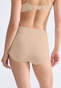 Kvinna i beige högmidjad sömlösunderkläder och matchande BH, sedd bakifrån mot en enkel ljus bakgrund.