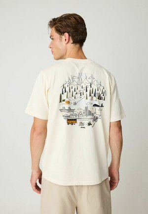 Uomo che indossa una maglietta color crema con illustrazione di paesaggio esterno e urbano che include montagne, alberi, tende, fiume, animali, veicoli e edifici.