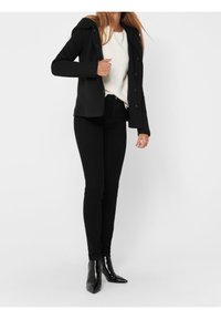 Svart figursydd jacka med huva, över en vit tröja, kombinerad med svarta slim fit jeans och spetsiga svarta ankle boots. Släta texturer och modern design.