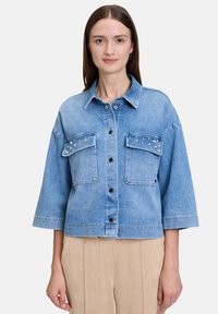Giacca di jeans blu chiaro con colletto, due tasche frontali decorate con borchie e bottoni neri. Vestibilità relaxed e cropped con maniche ampie.