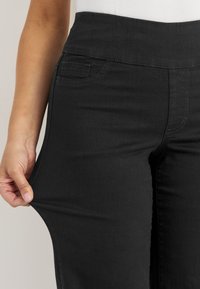 Henkilö venyttää mustia istuvia housuja, mikä näyttää kankaan joustavuuden ja korkean vyötärön yksityiskohdan.