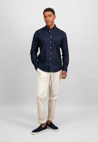 Marine skjorte med lange ermer, hvite knapper, beige chinos med oppbrett, slip-on marine blå sko med teksturert overdel, stående på en nøytral bakgrunn.