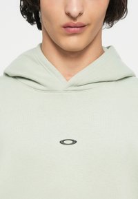 Ανοιχτό πράσινη φούτερ με κουκούλα που διαθέτει μπροστινή τσέπη καγκουρό, ρυθμιζόμενη κουκούλα με κορδόνι και μαύρο οβάλ λογότυπο στο στήθος. Μαλακό υλικό από μείγμα βαμβακιού.