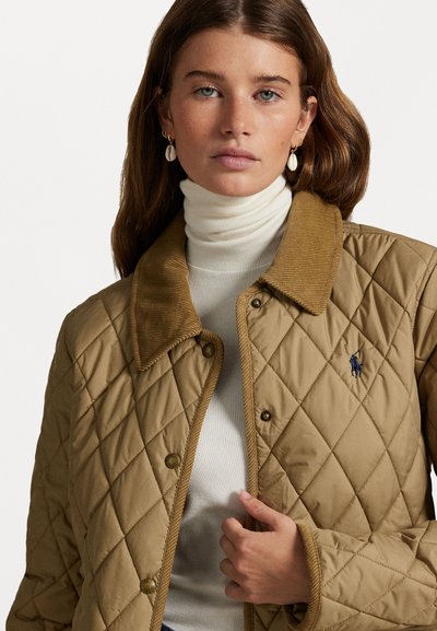 Veste matelassée beige avec un col en velours côtelé, dotée de boutons-pression et d'un logo brodé. Portée sur un pull col roulé blanc.