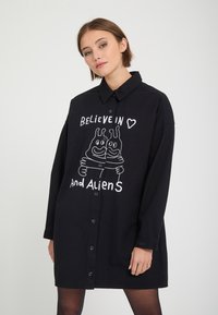 Jeune femme portant une robe-chemise noire boutonnée avec un texte blanc et un dessin de deux extraterrestres en train de s’enlacer et les mots "Believe in and aliens."