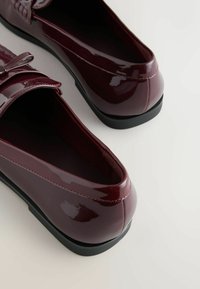 Bordeauxfarbene Lacklederschuhe mit glänzender Oberfläche, flacher schwarzer Sohle und gesteppten Akzenten am Obermaterial. Rund zulaufende Zehenform.
