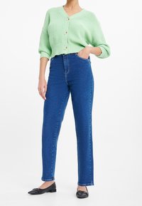 Osoba oblečená v modrých džínech s vysokým pasem, v světle zeleném zkráceném pleteném kardiganu s knoflíky a v  černých špičatých balerínách, stojí s jednou rukou v kapse.