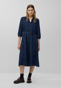 Denim shirtjurk in donkerblauw, met knopen en een strikceintuur, 3/4 mouwen en een knielange A-lijn rok. Draagt met donkere sokken en chunky schoenen.