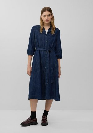 Robe chemise en denim bleu foncé, boutonnée avec une ceinture à nouer, manches 3/4 et jupe évasée à mi-genoux. Portée avec des chaussettes foncées et des chaussures épaisses.