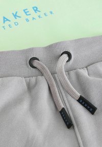 Grijze joggingsbroeken met een geribde tailleband en trekkoord, zwarte oogjes en logo-tags met de tekst "BAKER" in het blauw. Zacht, textuurmateriaal is zichtbaar.