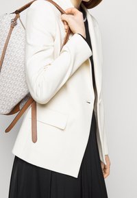 Hvid skræddersyet blazer med slim fit, udstyret med en enkelt lomme og udskårne revers, parret med en lysegrå mønstret rygsæk med brune stropper.