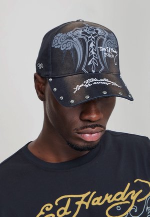 Black man wearing a black cap with a decorative cross and text, paired with a black t-shirt with gold "Ed Hardy" branding.