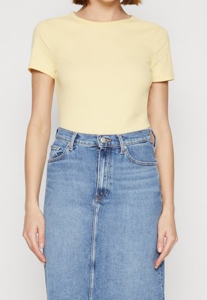 Femme portant un haut jaune côtelé à manches courtes, rentré dans une jupe en denim bleu taille haute avec fermeture à boutons et zip.