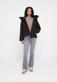 Giacca nera corta con colletto alto, realizzata in un tessuto texture. Indossata su un top beige, abbinata a jeans svasati blu chiaro e sneakers.