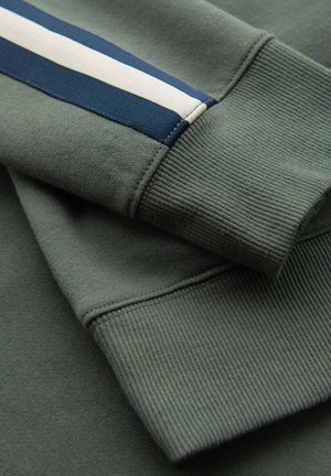 Opgevouwen mouwen van een olijfgroene trui met geribde boorden, met een blauw-wit gestreept detail op één mouw.