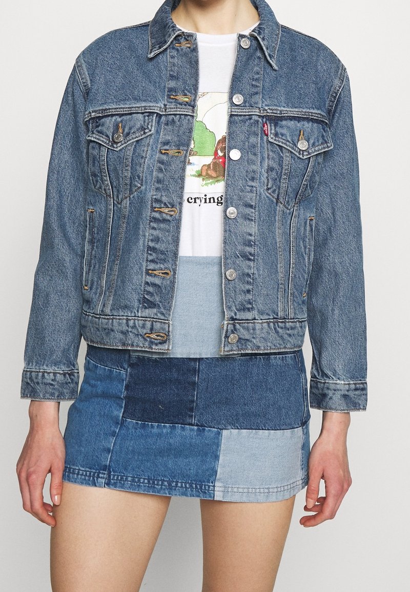 Personne portant une veste en denim bleu par-dessus un t-shirt blanc à motif graphique et une mini-jupe en denim patchwork avec différentes nuances de bleu.