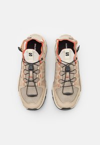 Des chaussures de sport beige avec un dessus en maille, des accents orange, des lacets élastiques et une semelle texturée. Conception légère pour une meilleure respirabilité.