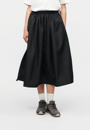 Femme portant une jupe midi noire à taille élastique, des chaussettes blanches, des chaussures de randonnée grises et un haut blanc à manches courtes, debout sur fond blanc.