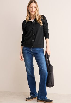 Suéter negro abotonado con dos bolsillos en el pecho, combinado con jeans azules de pata de elefante y zapatillas negras, sosteniendo un bolso negro.