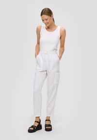 Weißes ärmelloses Tanktop kombiniert mit hellen weißen Leinen-Hosen mit Kordelzug und Taschen. Tragen mit schwarzen Plateausandalen. Minimalistisches Design.