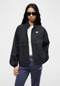 Γυναίκα με μακριά σκούρα μαλλιά φοράει μαύρο μπουφάν New Balance, λευκό crop top, ναυτικό παντελόνι φόρμας και μαύρα γυαλιά ηλίου, στέκεται σε λευκό φόντο.