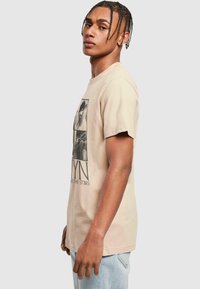 Beige bavlněné tričko s krátkým rukávem, s grafickým designem na přední straně. Model má na sobě světle modré džíny a stojí bokem.