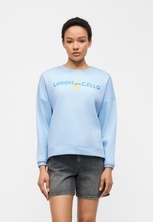 Γυναίκα που φοράει γαλάζιο φούτερ με κείμενο «Limoncello» και γραφικό με λεμόνι, σε συνδυασμό με μαύρο τζιν σορτς, στέκεται μπροστά σε λευκό φόντο.