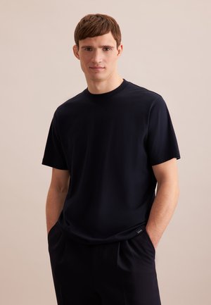 Jeune homme aux cheveux courts et bruns portant un t-shirt noir uni à manches courtes et un pantalon noir, debout les mains dans les poches devant un fond uni.