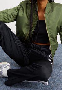Giacca bomber in satin verde, top corto nero, pantaloni sportivi neri con strisce bianche e scarpe da ginnastica grigie con accenti bianchi.