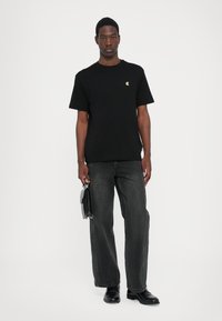 Tricou negru din bumbac, cu mâneci scurte și un mic logo auriu, asortat cu blugi largi de culoare gri închis și pantofi negri, purtând o geantă neagră.