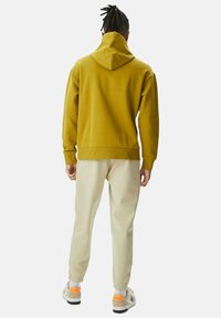 Sudadera con capucha color mostaza y corte relajado, combinada con pantalones de chándal beige claros y zapatillas blancas con detalles en naranja.