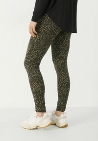 Leggings verts avec un motif à pois noirs, tissu extensible et coupe slim, associés à des baskets blanches avec des accents beiges.