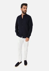Camicia a maniche lunghe blu navy con colletto rialzato, chiusura con bottoni e design aderente, abbinata a pantaloni bianchi e espadrillas nere.