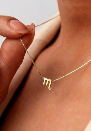 Hand die een gouden ketting met een gouden Scorpio sterrenbeeldhanger vasthoudt, gedragen op blote huid bij het sleutelbeen.