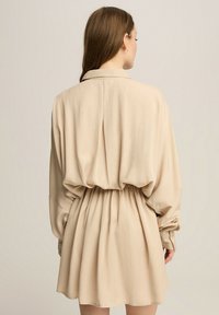 Beżowa sukienka koszulowa z kołnierzykiem, długimi rękawami i elastycznym paskiem. Miękka tkanina, luźny krój i rozkloszowany dół. Gładka faktura.