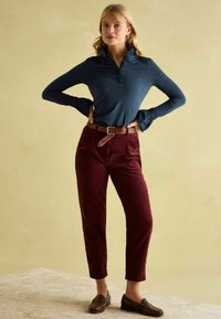 Gestreiftes, navyblaues Langarm-Polohemd, kastanienbraune Cordhosen mit einem gewebten braunen Gürtel, braune Slipper, vor einem hellen Hintergrund.