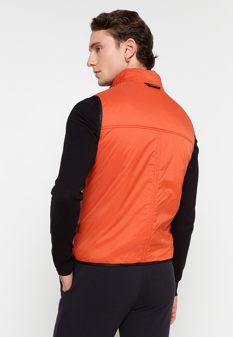 Mężczyzna w jaskrawopomarańczowej kamizelce bez rękawów na czarnej koszuli z długim rękawem, stojący z rękami w kieszeniach, zerkający w lewo.