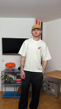 Ung mann med tatoveringer som har på seg en beige caps og hvit Adidas-skjorte, står i et rom med TV, oransje lampe, planter og en klistret koffert.